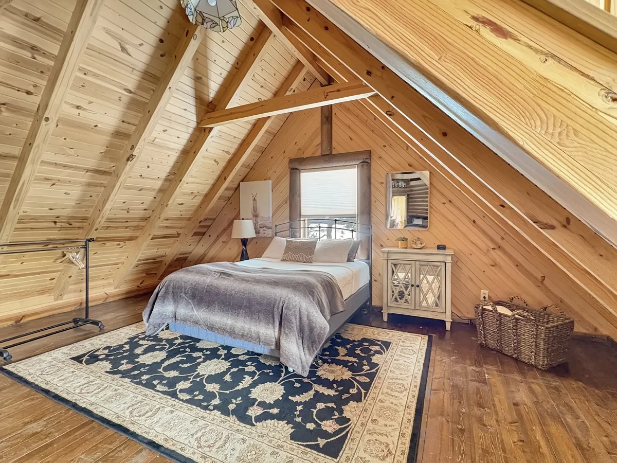 A cozy attic bedroom with wood-paneled slanted walls and ceiling, featuring a bed with pillows and a blanket, a nightstand with a lamp, a mirror, a basket, and a decorative rug on the wooden floor.
