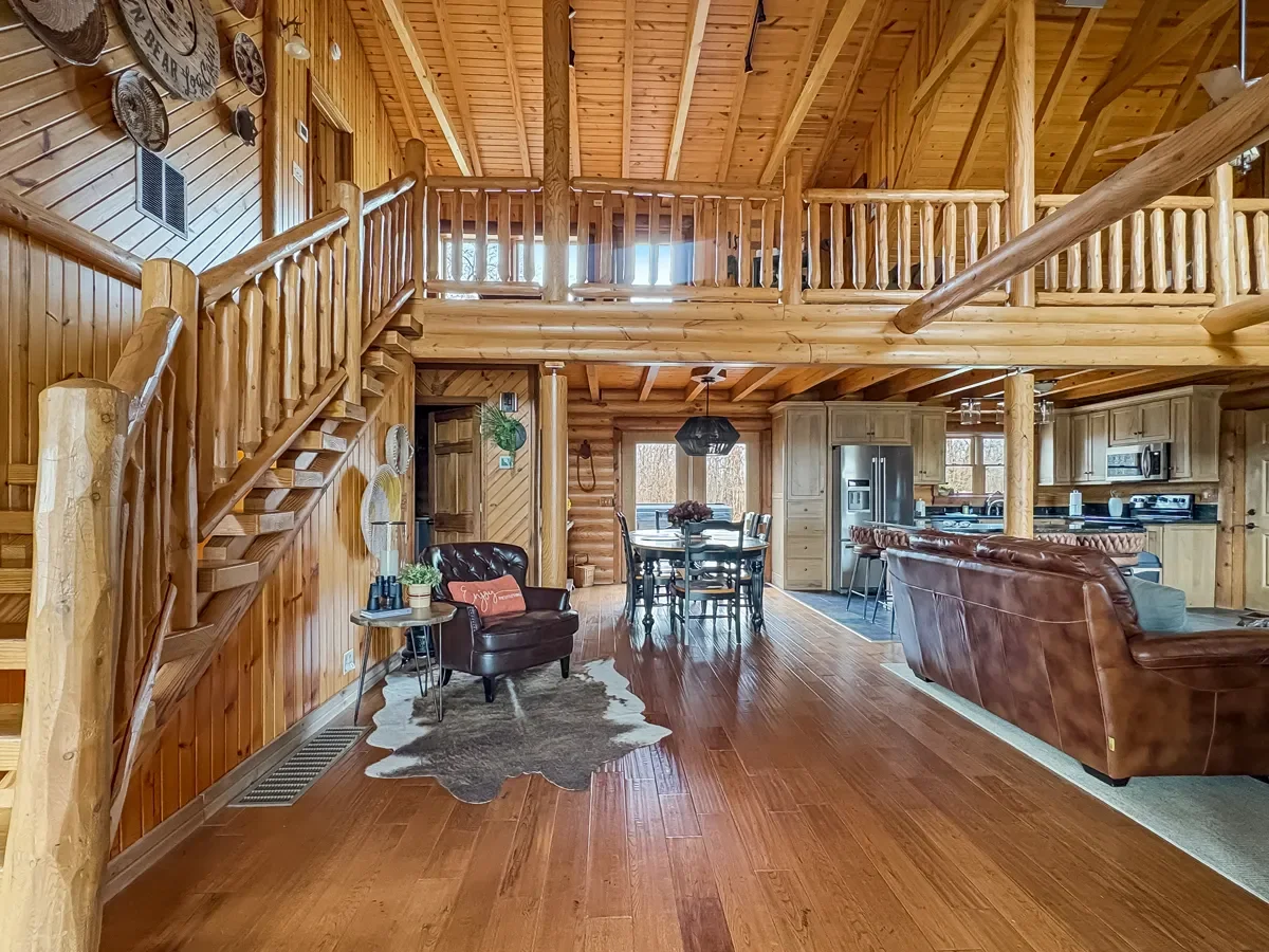Interior of a log cabin with wooden walls and ceiling, featuring a staircase with a wooden railing, a living area with leather sofas, a dining table with chairs, and a kitchen with wood cabinets and stainless steel appliances.