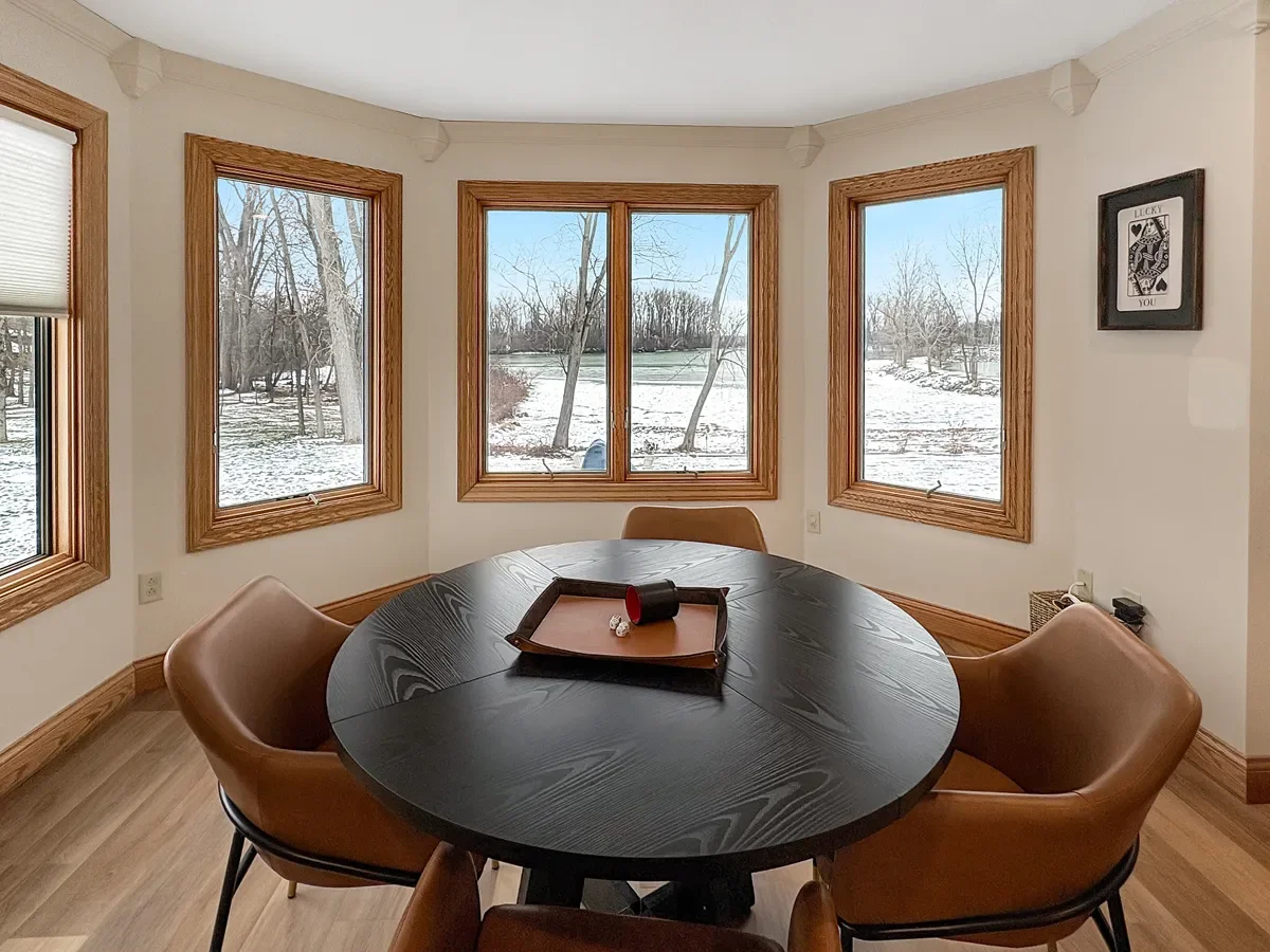 Dining room with round black table and four brown chairs, large windows showing snowy landscape with trees, wall decor with playing card picture.