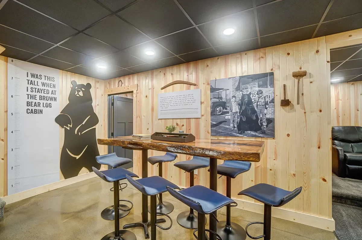 A cozy room with wood-paneled walls, black ceiling tiles, and a high wooden table with six stools. On one wall, there's a large bear silhouette with height measurements and text. The wall also features framed artwork and small decor items.