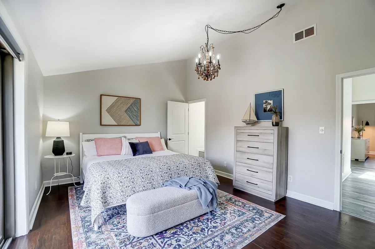 A bedroom with a white bed, pink and navy pillows, a patterned bedspread, a gray dresser with decorative items, a chandelier, and a colorful area rug.