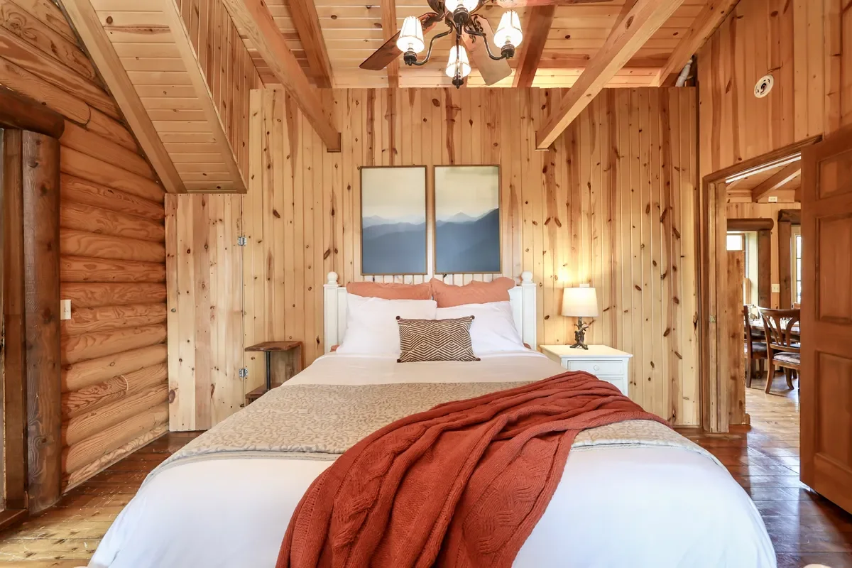 A cozy bedroom with wood-paneled walls, a white bed with orange and white bedding, a throw blanket, two bedside tables with lamps, and wall art depicting mountains.