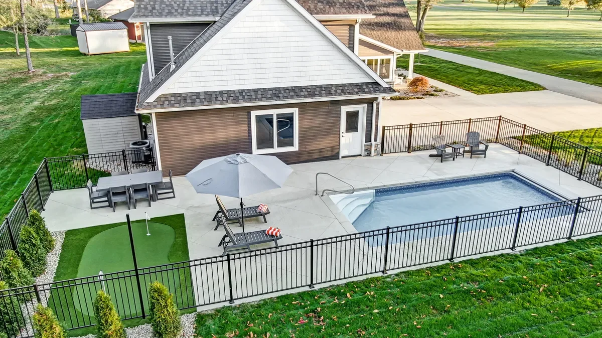 A backyard with a small swimming pool, lounge chairs with umbrellas, an outdoor dining table, and a mini golf putting green, all enclosed by a black metal fence, next to a house with a brown and white exterior.