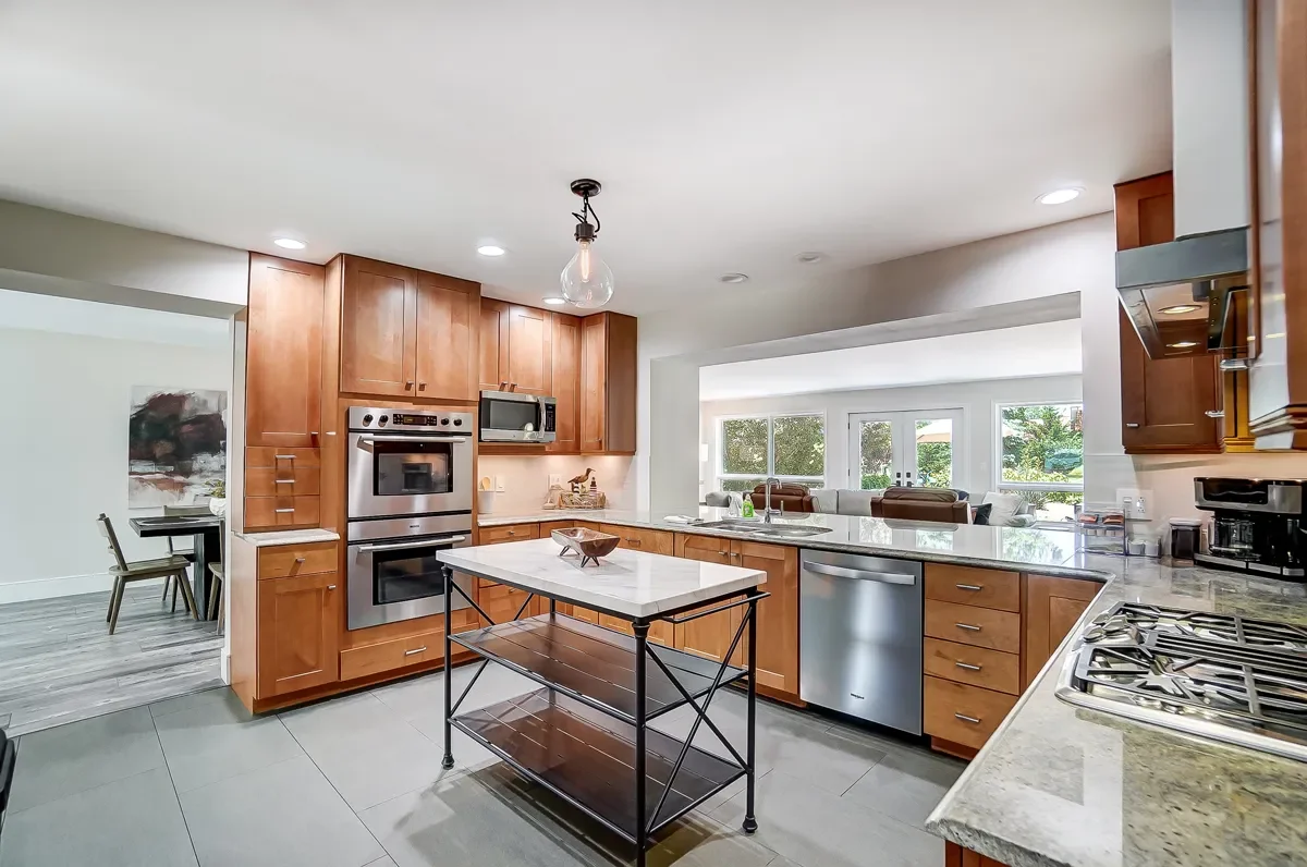 Modern kitchen with wooden cabinets, stainless steel appliances, white marble island, and a view into the dining and living areas.