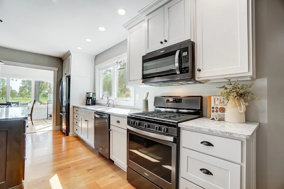 Kitchen with white cabinets, stainless steel appliances including microwave and oven, window above sink, wooden flooring, and a small decorative plant on the right