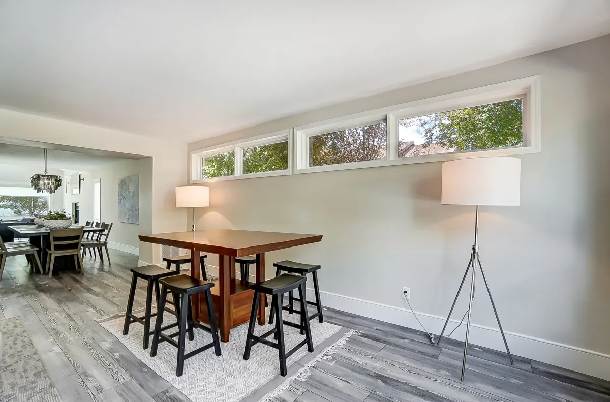 Modern dining room with a high window, wooden table, black stools, and two floor lamps, with a view of an adjacent dining area.