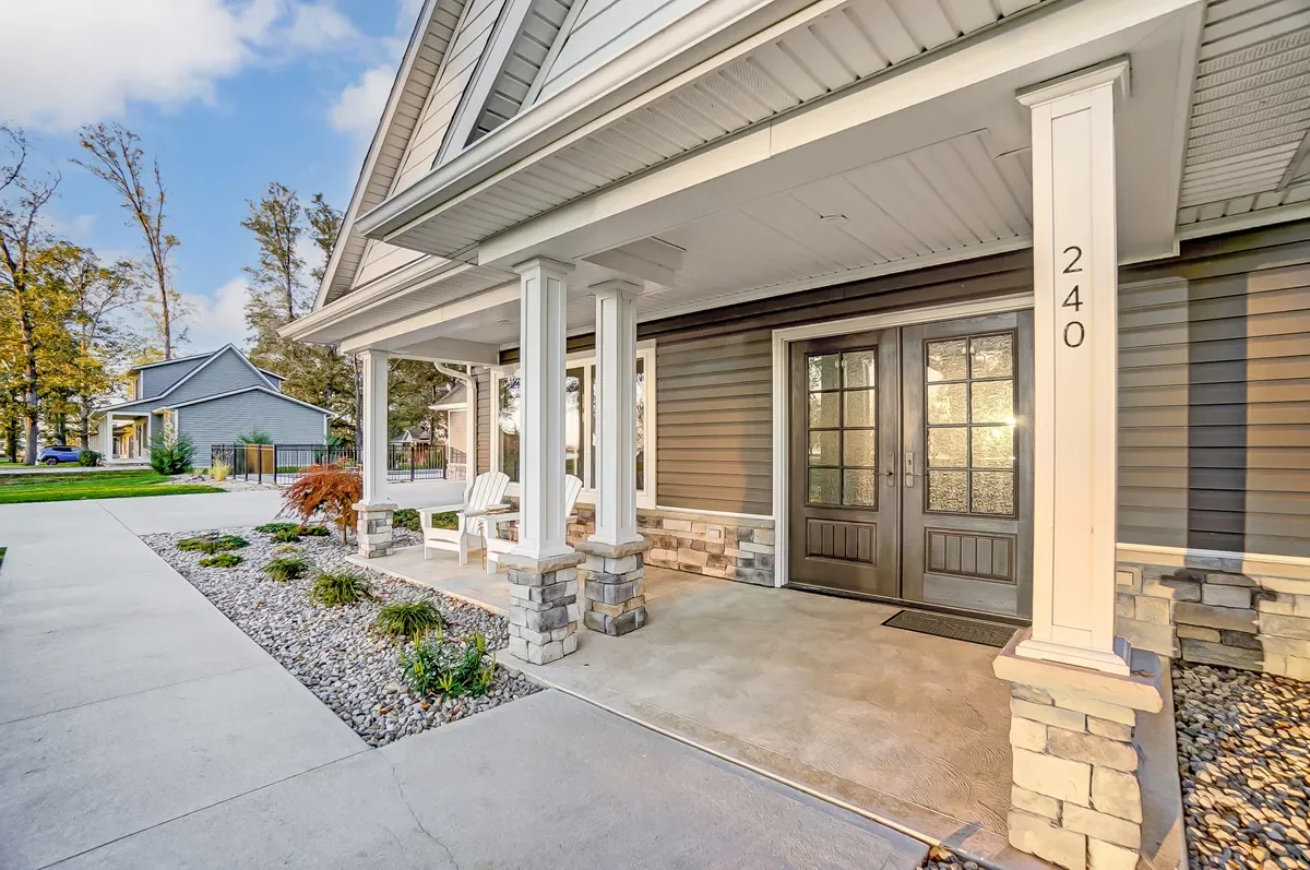 Front porch of a modern house with a covered entryway, white pillars, stone bases, gray siding, double black front doors, and a concrete walkway surrounded by landscaped garden beds with rocks and plants.