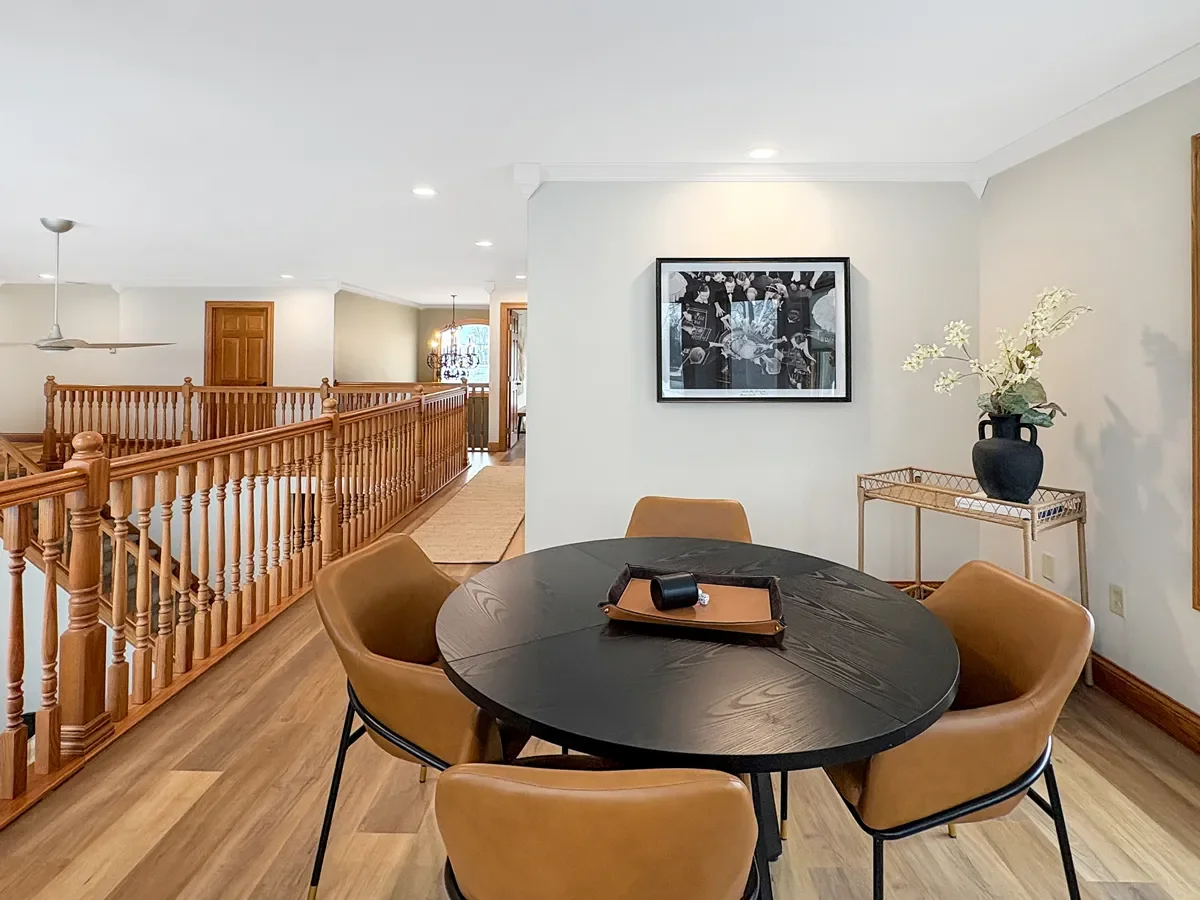 Interior view of a dining area with a black round table, four tan chairs, a tray with a candle, a wall-mounted picture, a side table with a black vase and flowers, and a wooden railing in the background.