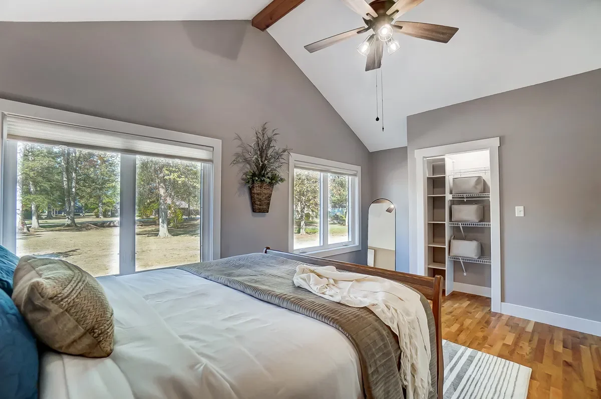 A cozy bedroom with a vaulted ceiling, a ceiling fan, three large windows showing an outdoor scene with trees, a bed with pillows and blankets, a full-length mirror, and an open closet with shelves and storage bins.