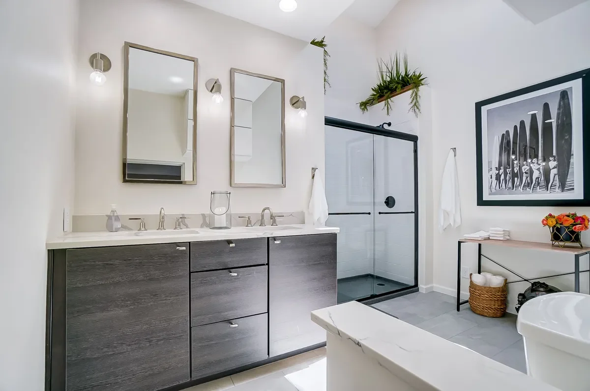 Modern bathroom with double vanity, two mirrors, wall-mounted lights, glass shower with black frame, framed artwork, and decorative plants.