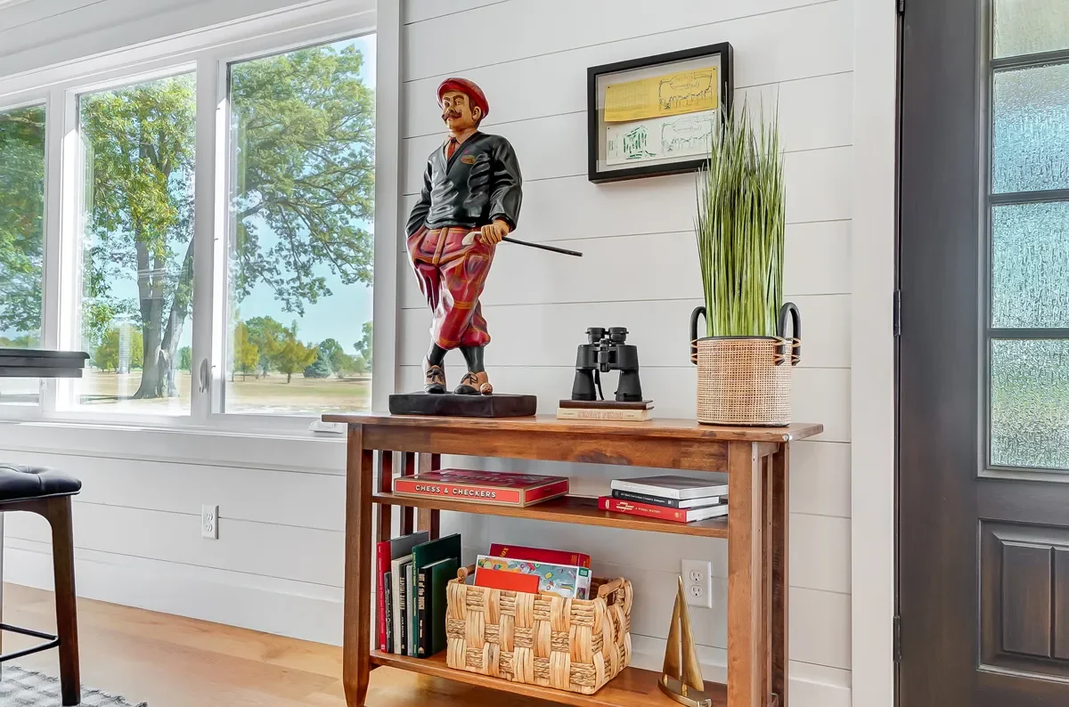 A wooden console table inside a house with decorative items, including a painted figure of a man, a pair of binoculars, a potted plant, and books, positioned near large windows revealing trees and greenery outside.