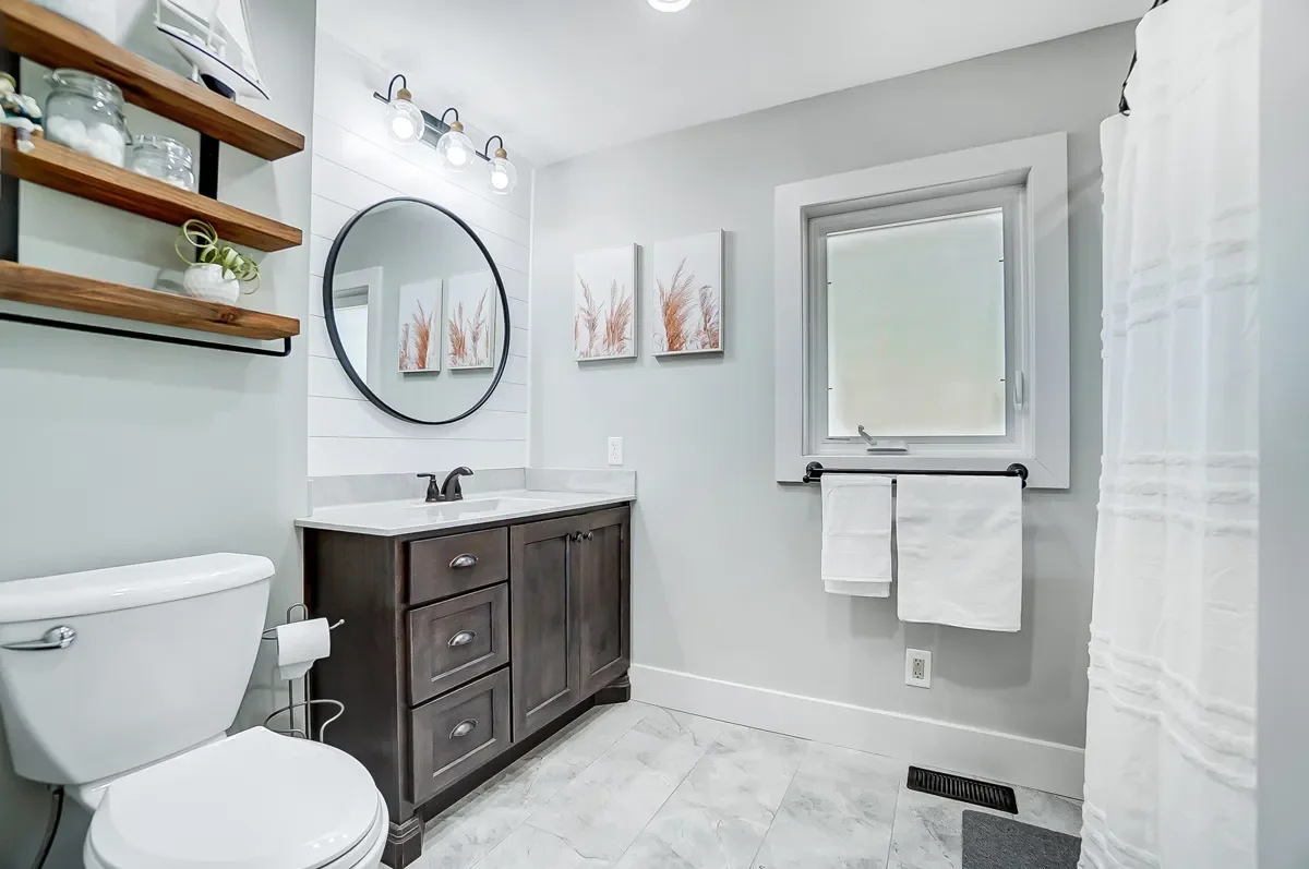 Modern bathroom with gray walls, a dark wooden vanity with a white countertop, a round mirror, artwork with botanical prints, white towels on a black towel bar, a window, and a white shower curtain.