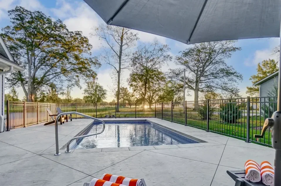 Quiet backyard swimming pool with a safety fence, lounge chairs, large umbrella, and trees in the background during sunset.