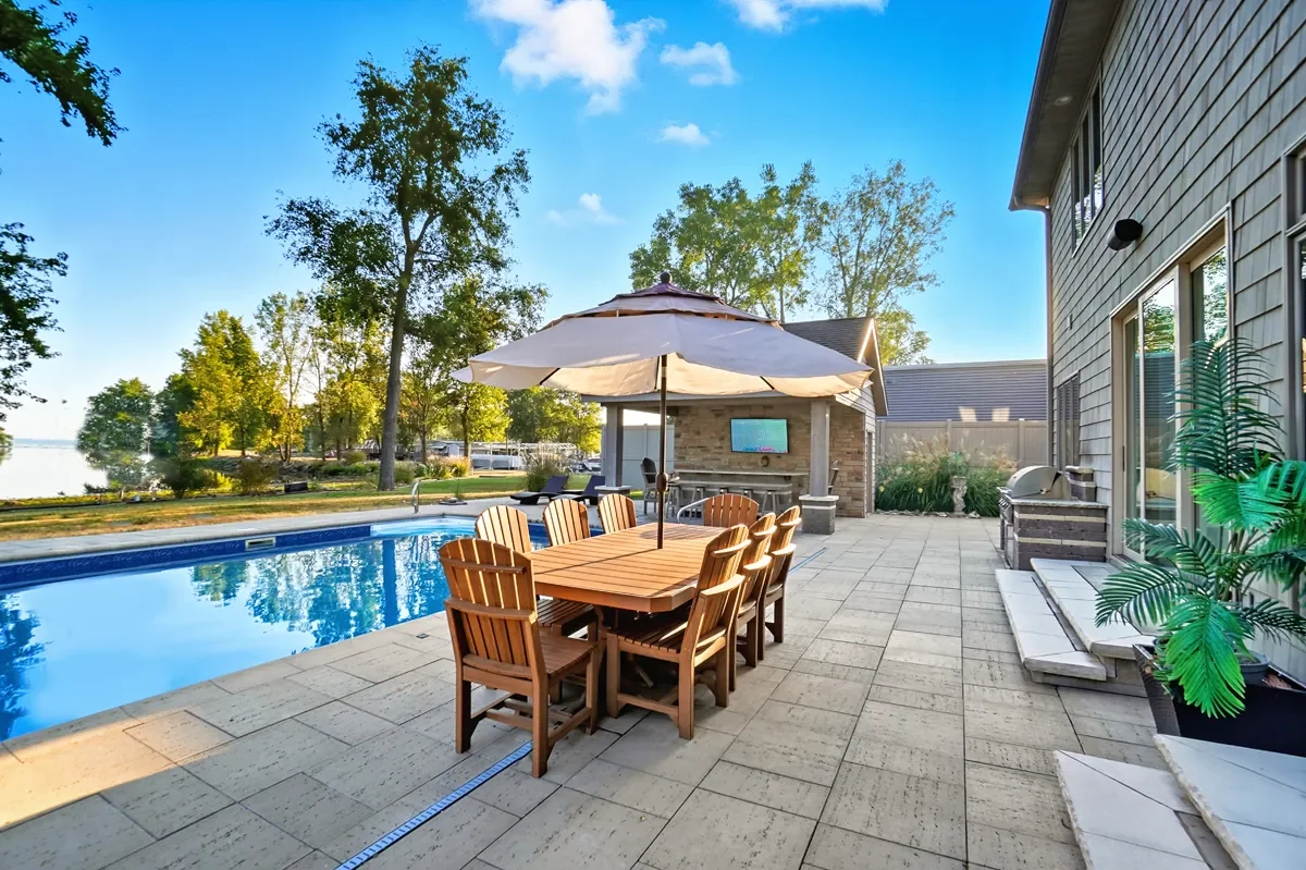 A backyard with a swimming pool, outdoor dining table with chairs and an umbrella, trees, and a building with large windows on a sunny day.