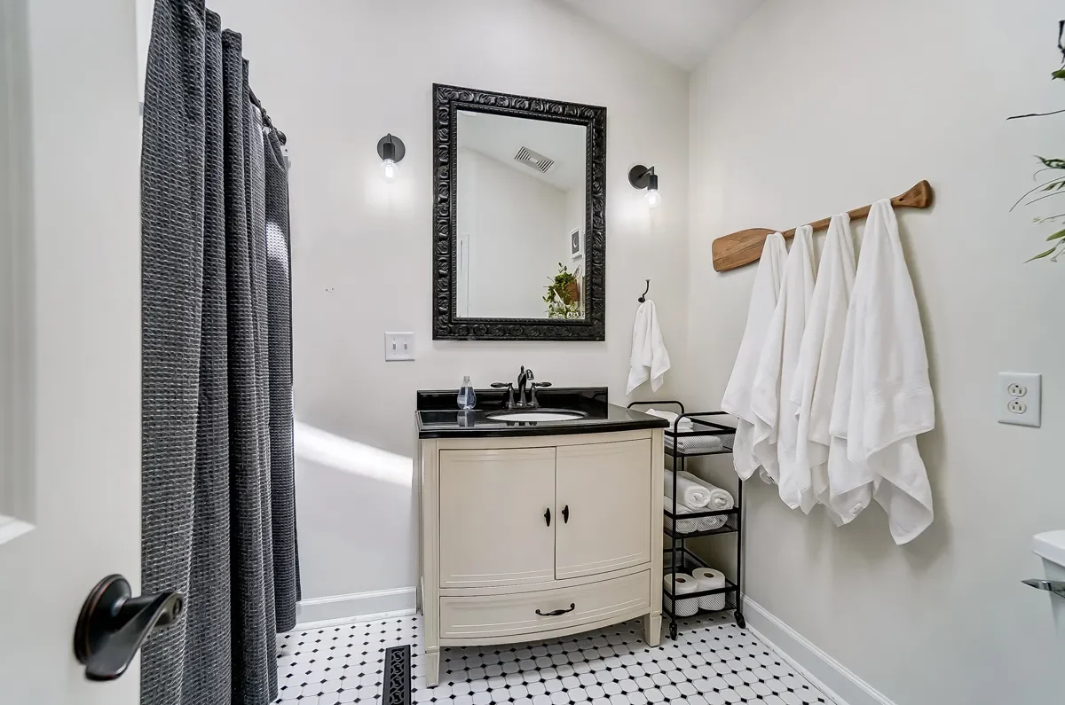 Bathroom with white walls, a black framed mirror above a cream vanity with black countertop, white towels hanging on a wooden rack, a black rolling towel shelf with white towels, and a black and white hex tile floor.