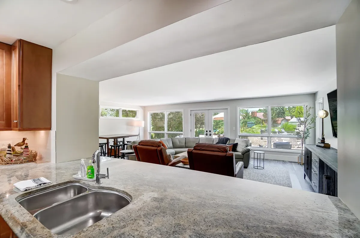 Open-concept living room with large windows, sectional sofa, armchairs, TV, and balcony. Kitchen with granite countertop in foreground.