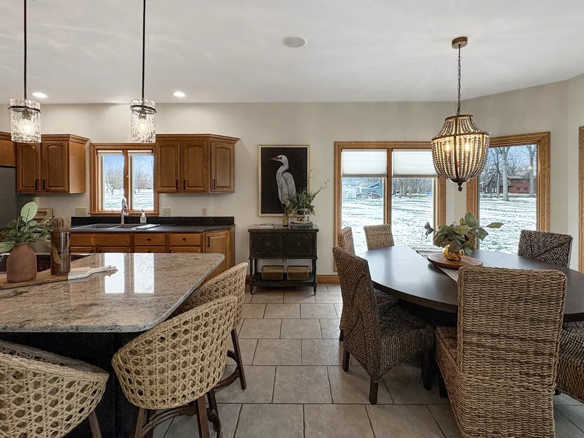 A dining area and kitchen with wooden cabinets, granite countertops, wicker chairs, a chandelier, large windows showing a snowy yard outdoors, and a framed bird painting on the wall.