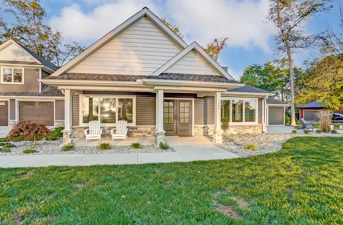 Front view of a modern house with a porch, two white chairs, and a small table, surrounded by a grassy lawn and trees.