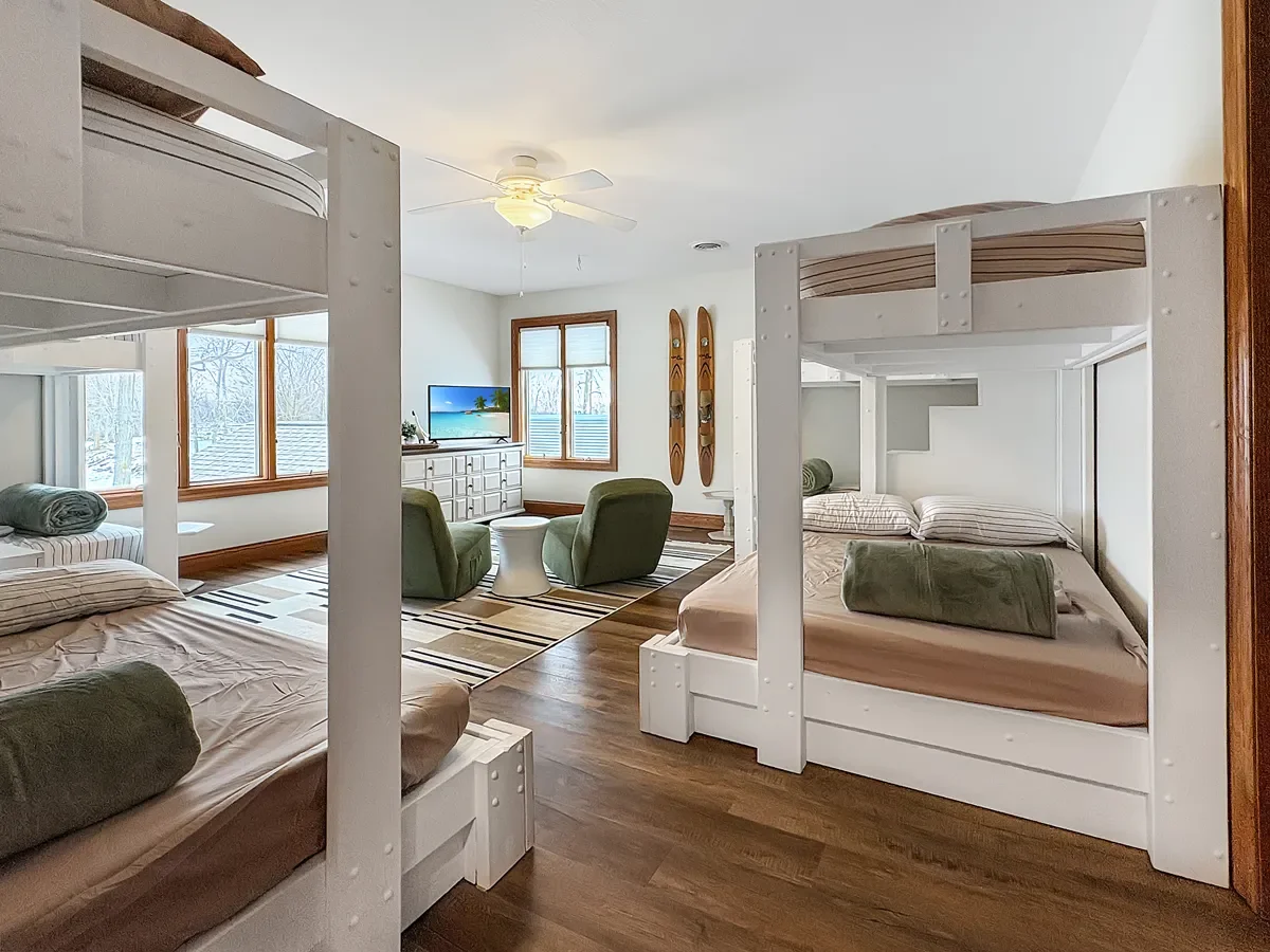 A cozy, bright room with two sets of bunk beds, a sitting area with two green armchairs, a small white side table, a window view of snow outside, a vanishing white dresser, a mounted flat-screen TV, and decorative skis on the wall.
