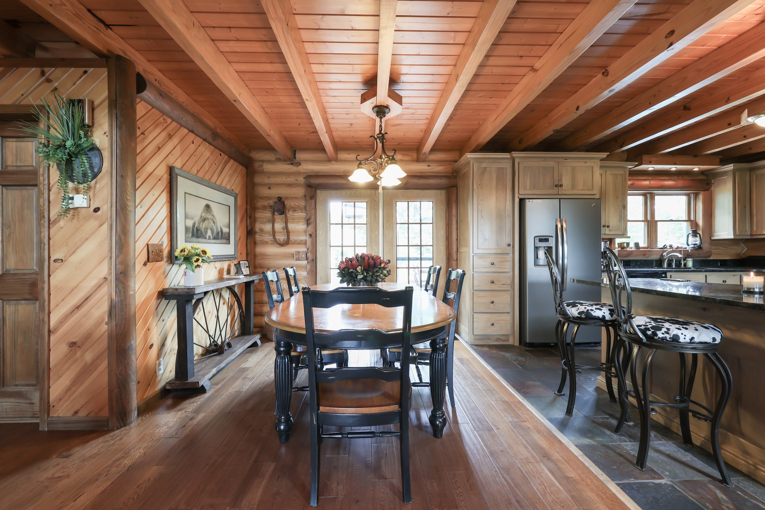 Rustic kitchen and dining area with wooden walls and ceiling, featuring a wooden dining table with black chairs, a stainless-steel refrigerator, bar stools, and a floral centerpiece.
