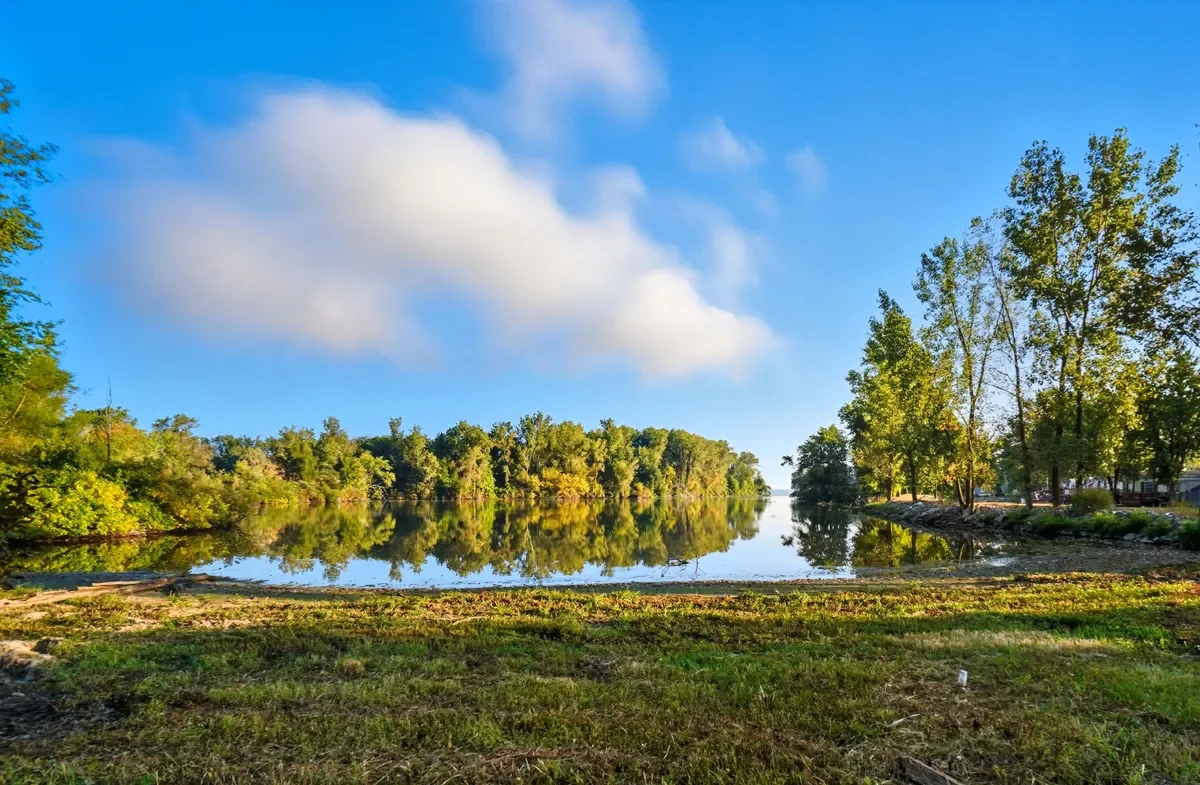 A peaceful river scene on a sunny day, with a calm river reflecting trees on both banks and a partly cloudy blue sky.