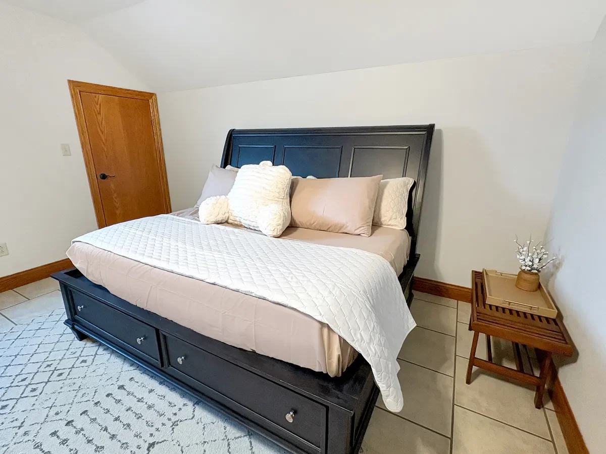 A bedroom with a black bed frame, beige bedding, and decorative pillows. There's a wooden door and a small wooden side table with a flower arrangement.