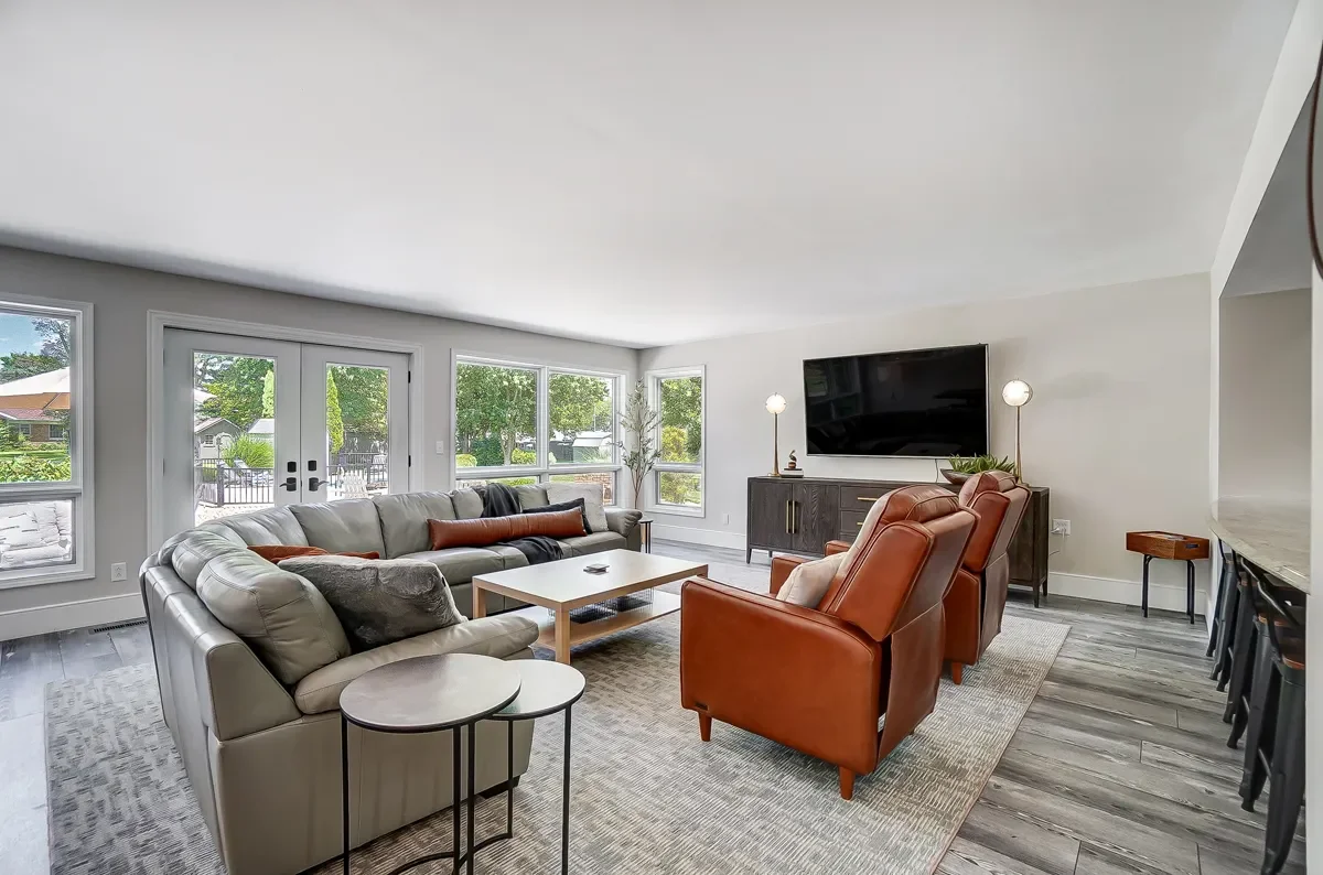 Bright living room with large windows, beige sofa, two orange armchairs, a black media console, mounted TV, and a rug on wooden flooring.