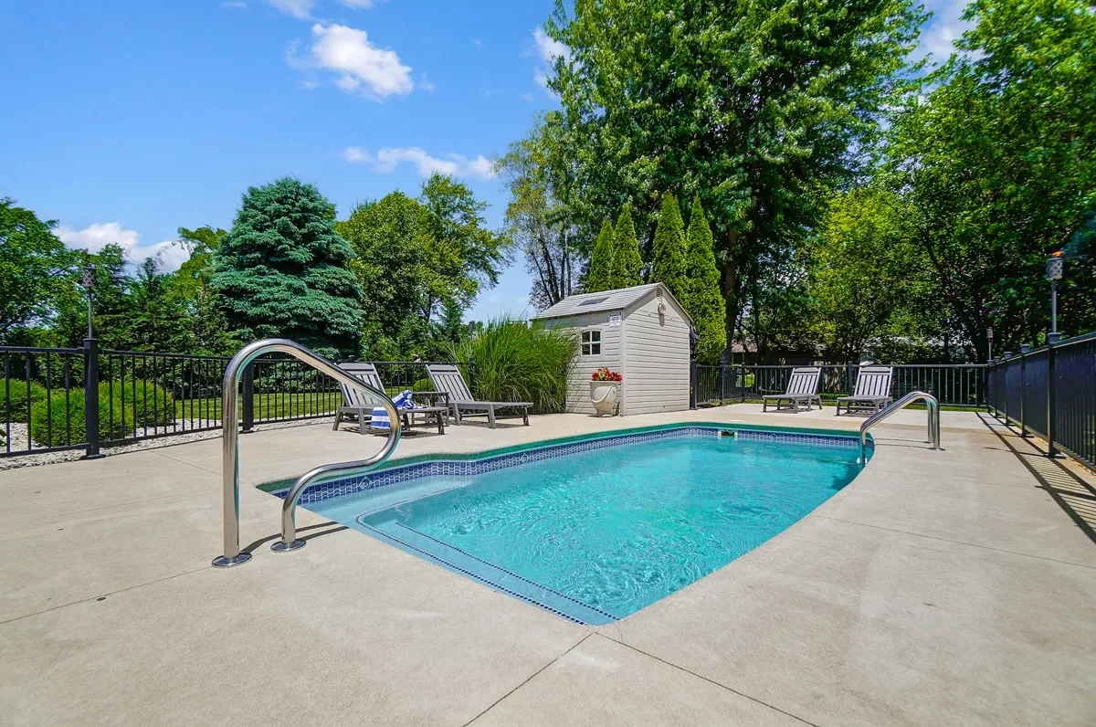 An outdoor swimming pool surrounded by a concrete deck, with lounge chairs, a small shed, and lush green trees in the background under a blue sky with some clouds.