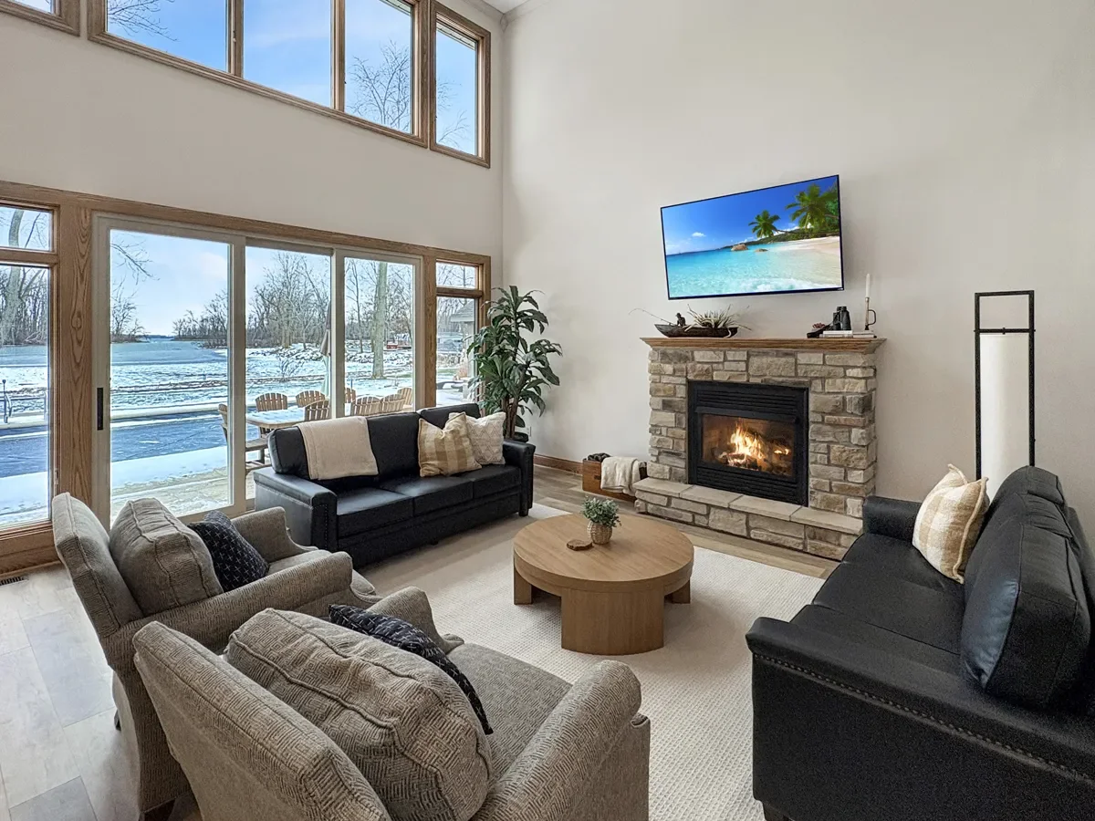 Living room with large windows showing a snowy outdoor scene, a fireplace, a flat-screen TV displaying a beach scene, and various seating furniture including sofas and armchairs.