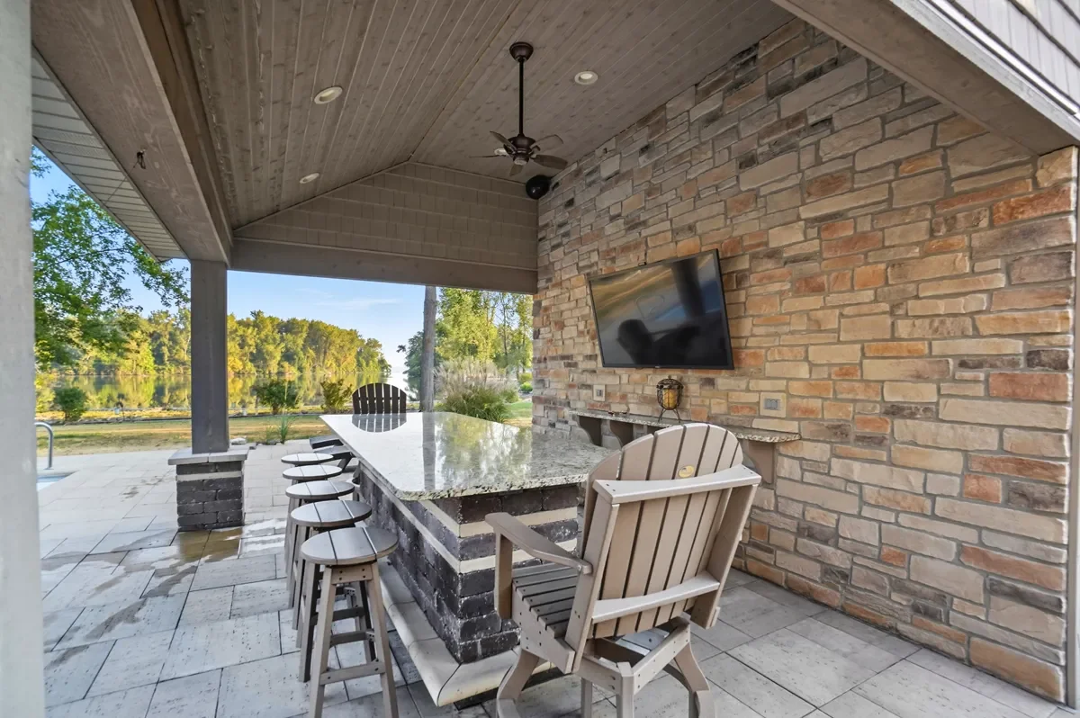 Outdoor covered patio with a high top granite bar, bar stools, a wooden armchair, a brick wall with a mounted TV, and a scenic view of a lake and trees.