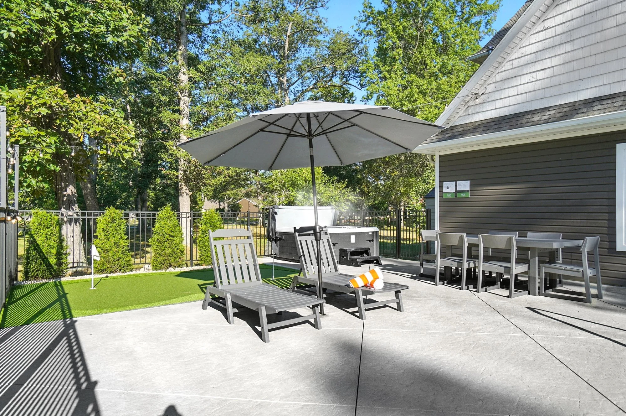 Backyard patio with lounge chairs, a large umbrella, a grill, and outdoor dining table with chairs. Green lawn area with trees and bushes in the background.
