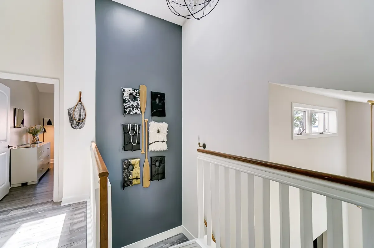 Interior view of a house staircase landing with a dark gray accent wall decorated with hanging fabric and leather pouches, a white faux fur bag, and a wooden paddle. There is a small window and a modern black metal chandelier hanging from the ceiling