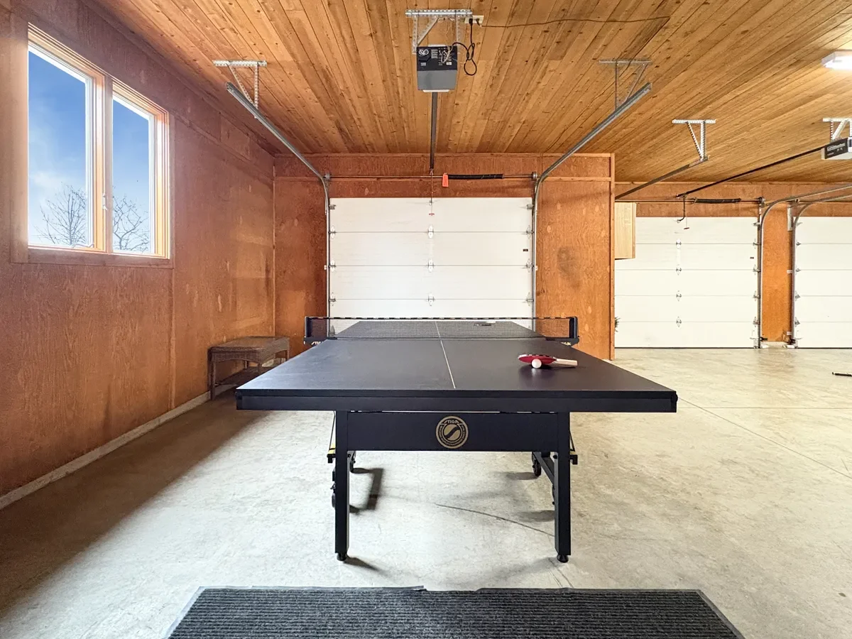 An indoor garage converted into a game room with a ping pong table, a ping pong paddle, and a ball on it. The garage has wood-paneled walls and ceiling, two overhead garage door openers, and a window letting in natural light.