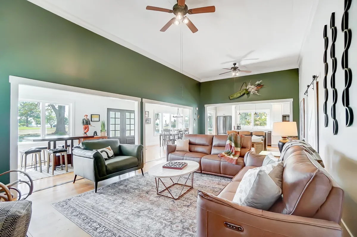 A cozy living room with green accent walls, leather sofas, a patterned area rug, a white coffee table, and large windows bringing in natural light, with a view of an outdoor area.