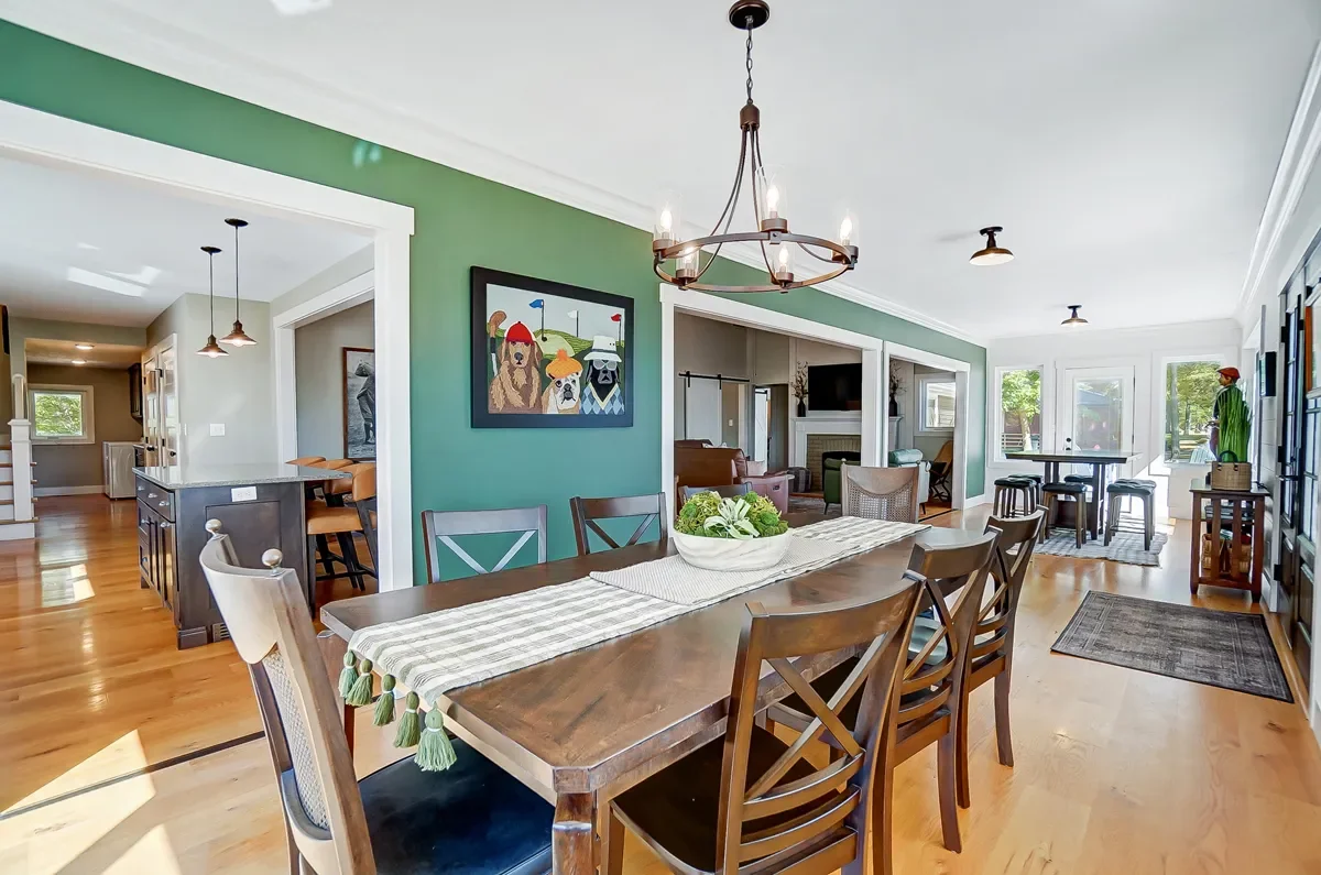 Open-concept dining and living area with hardwood floors, green accent wall, wooden dining table with six chairs, chandelier, and a large window with natural light.