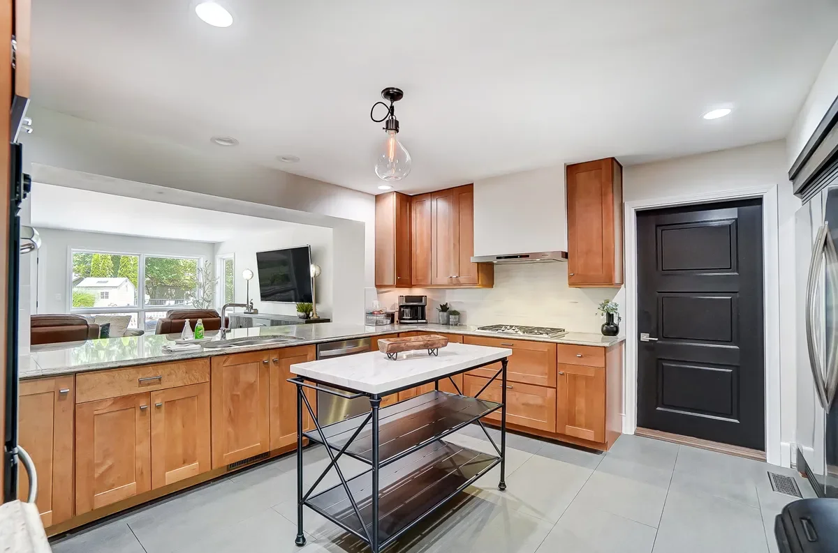 Modern kitchen with wooden cabinets, black door, marble island, and open view to living room with large window.