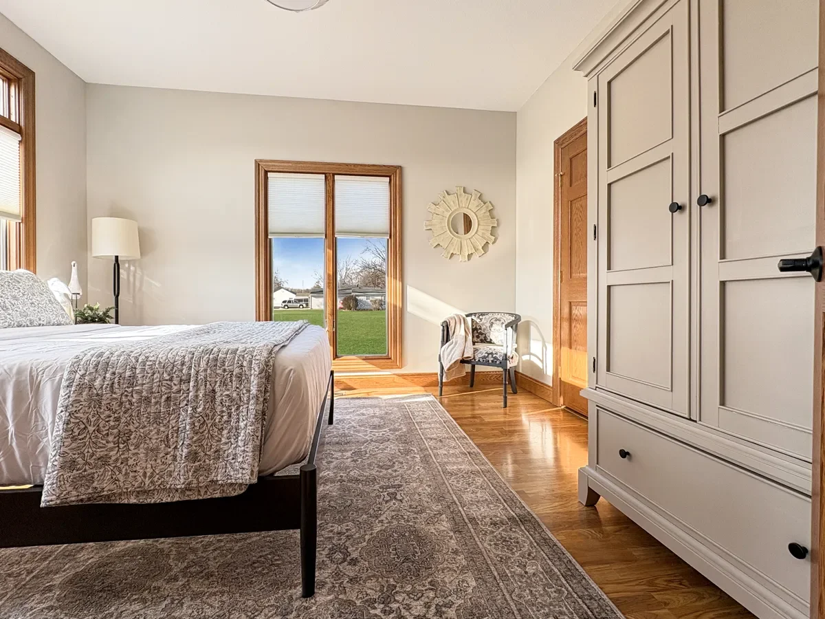 A bedroom with a bed, a window, a sliding glass door leading outside, a chair, a mirror, and a large wardrobe. Natural light illuminates the room.