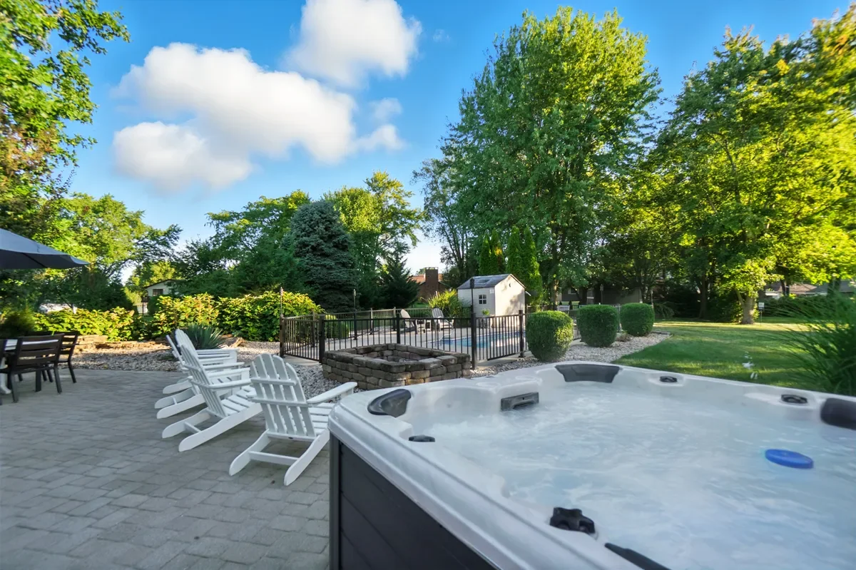  backyard scene with a hot tub in the foreground, patio chairs, and a fenced swimming pool, surrounded by lush green trees and shrubs under a blue sky with white clouds