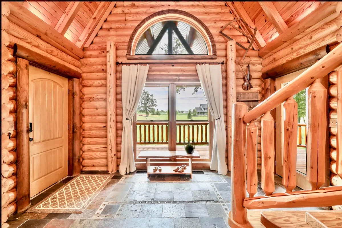 Interior of a log cabin with a wooden door, a large window with white curtains, and a view of a porch and grassy landscape outside. Rustic decor includes a violin and a saw hanging on the wall, with a small bench and potted plant near the window.