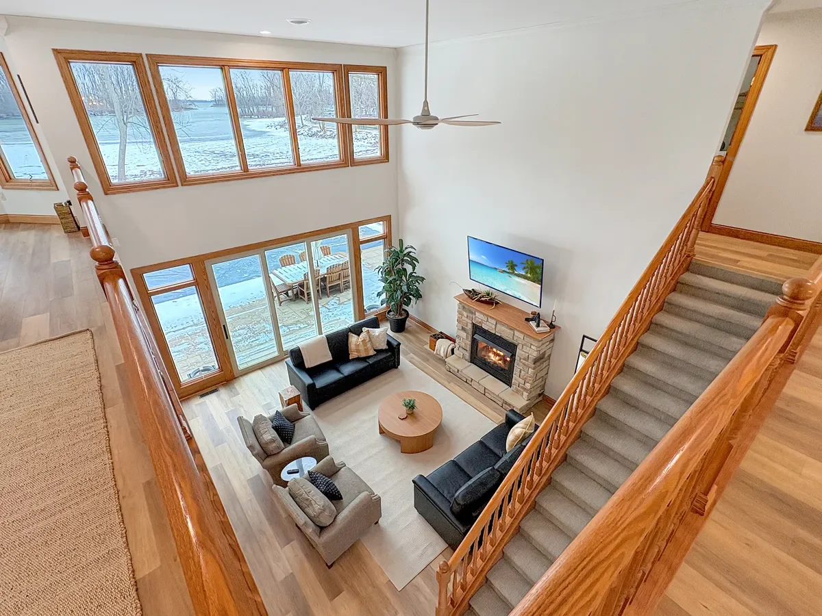 Living room with large windows overlooking snowy landscape, furnished with sofas, armchairs, a round coffee table, and a fireplace with a TV above, viewed from the staircase.
