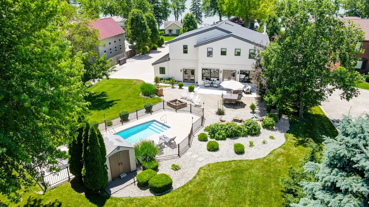 A backyard with a swimming pool surrounded by a black fence, a patio area with outdoor furniture, and a white modern house. The yard has well-maintained green grass and trees providing shade.