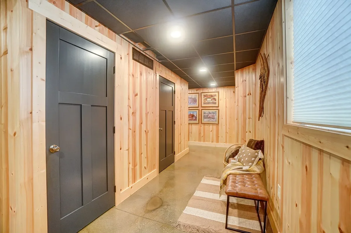 A hallway with light wood-paneled walls, gray doors, a black ceiling, and a concrete floor. There is a brown bench with cushions and a blanket, a striped rug, and framed pictures on the far wall.