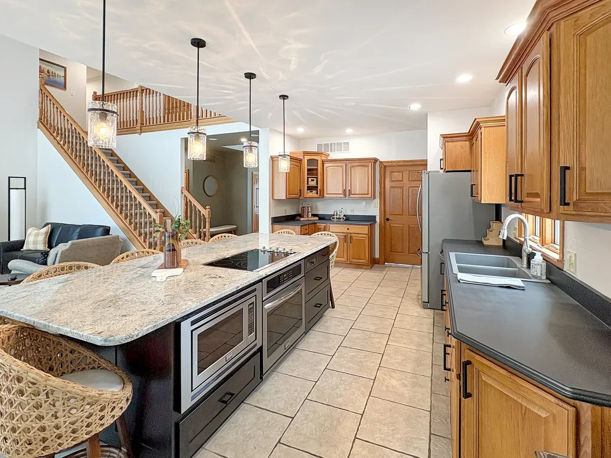 Open kitchen with wooden cabinets, black countertops, stainless steel appliances, and a kitchen island with a built-in stove, overlooking a living room with stairs and seating area.