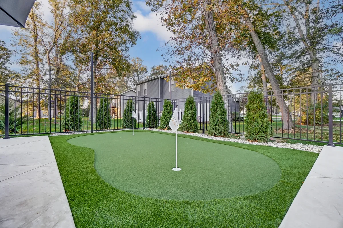 Backyard putting green with artificial turf, two white flags, surrounded by a black metal fence, trees, and a neighboring house in the background.