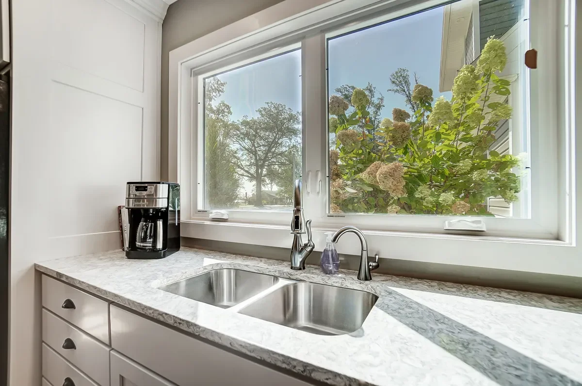 Kitchen counter with a coffee maker, double sink, and a window overlooking a garden with hydrangea bushes and trees.