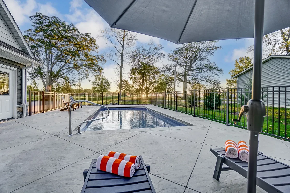 Resort-style backyard with a small swimming pool, sun loungers with rolled towels, large patio umbrella, and a view of trees and grass at sunset.