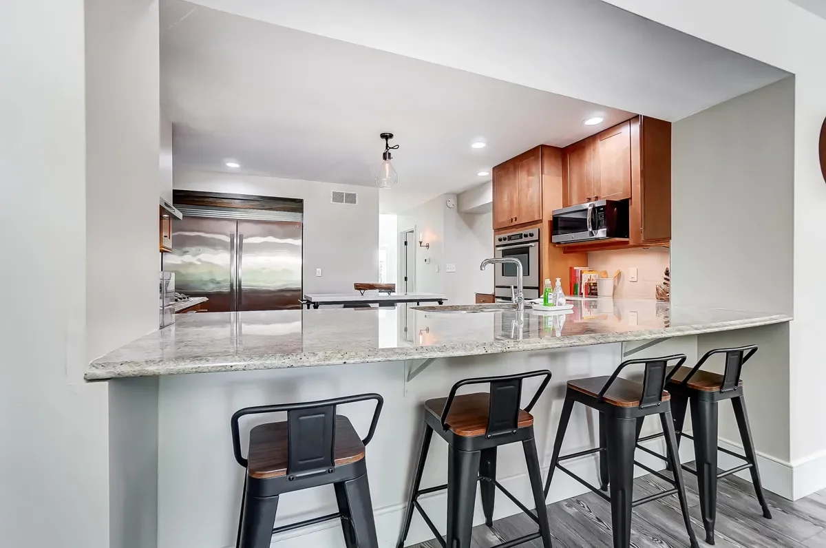 Modern kitchen with granite counter island, four bar stools, stainless steel refrigerator, microwave, oven, and wooden cabinets.
