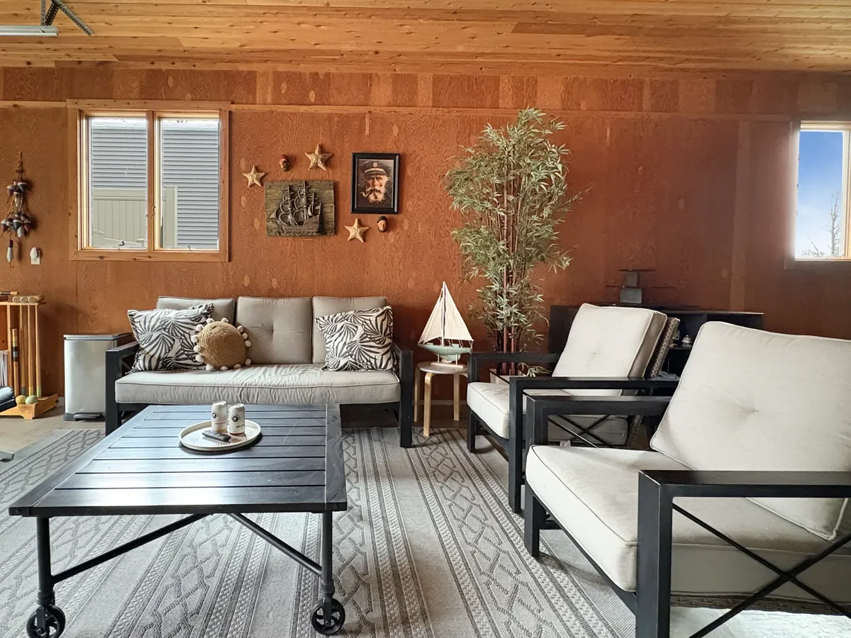Living room with wood-paneled walls, white sofa with decorative pillows, potted plant, wooden coffee table with candles, wall art, and windows with outdoor view.