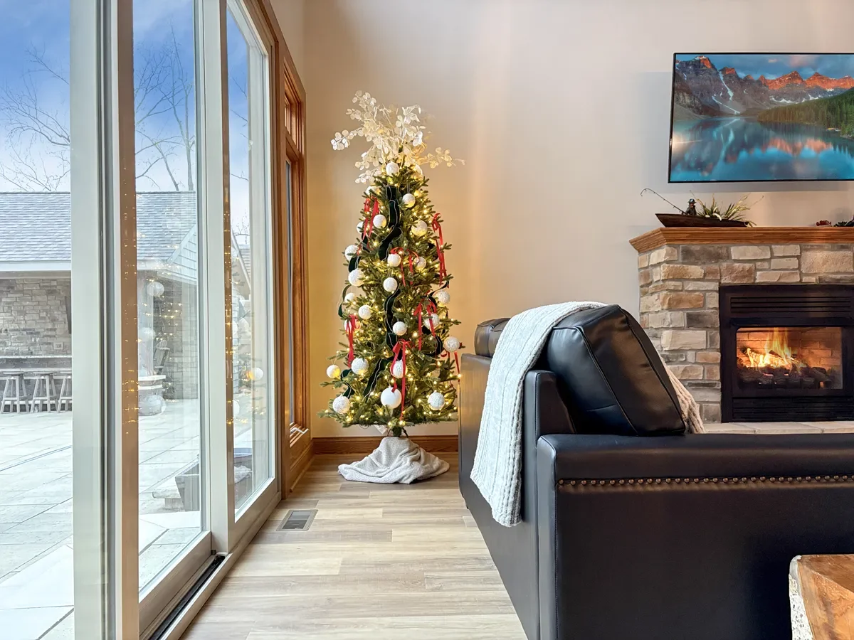 Decorated Christmas tree next to sliding glass doors in a living room with a fireplace and mounted TV.