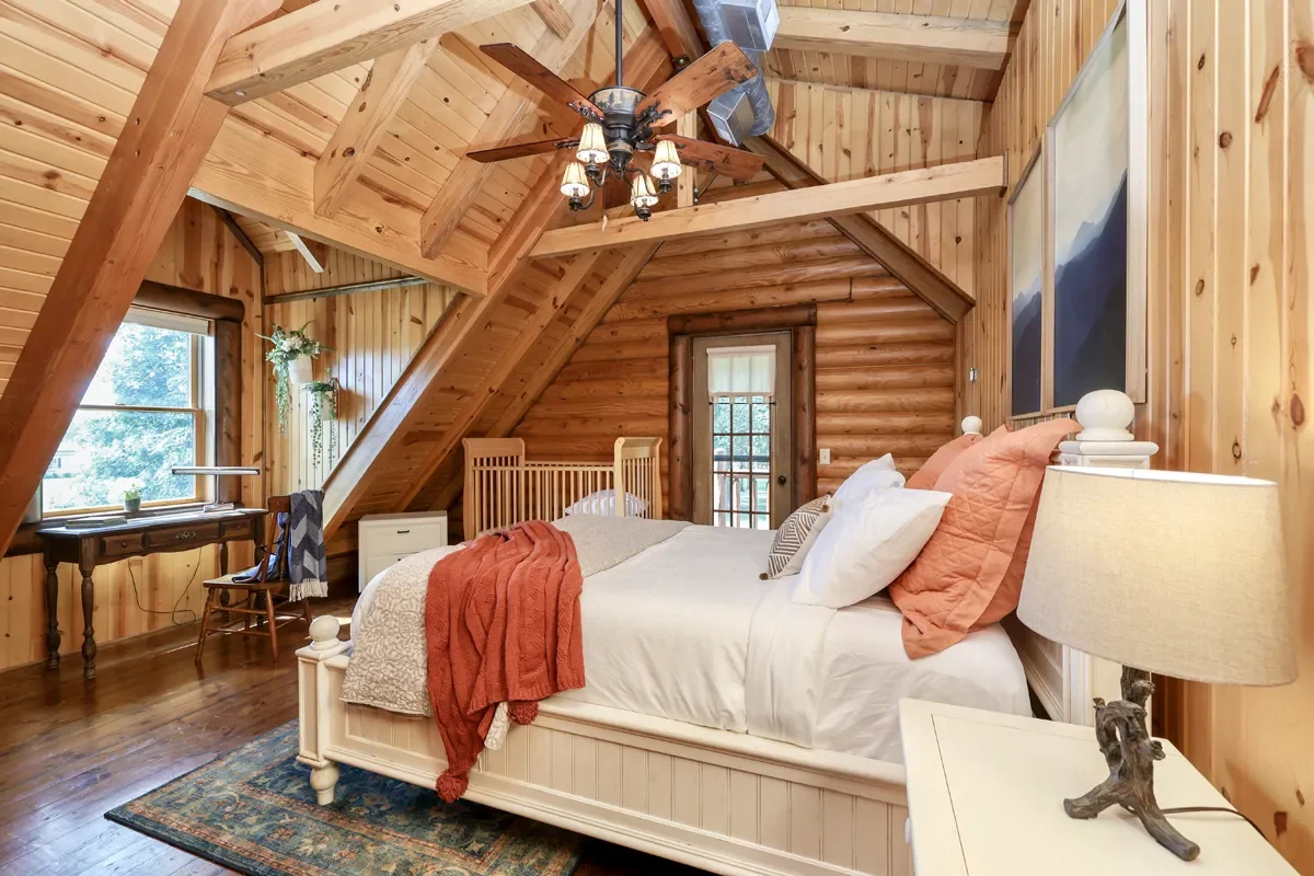 A cozy rustic bedroom with wood-paneled walls and ceiling, a bed with white and orange bedding, a wooden nightstand with a lamp, a window with a curtain, and a ceiling fan with light fixtures.